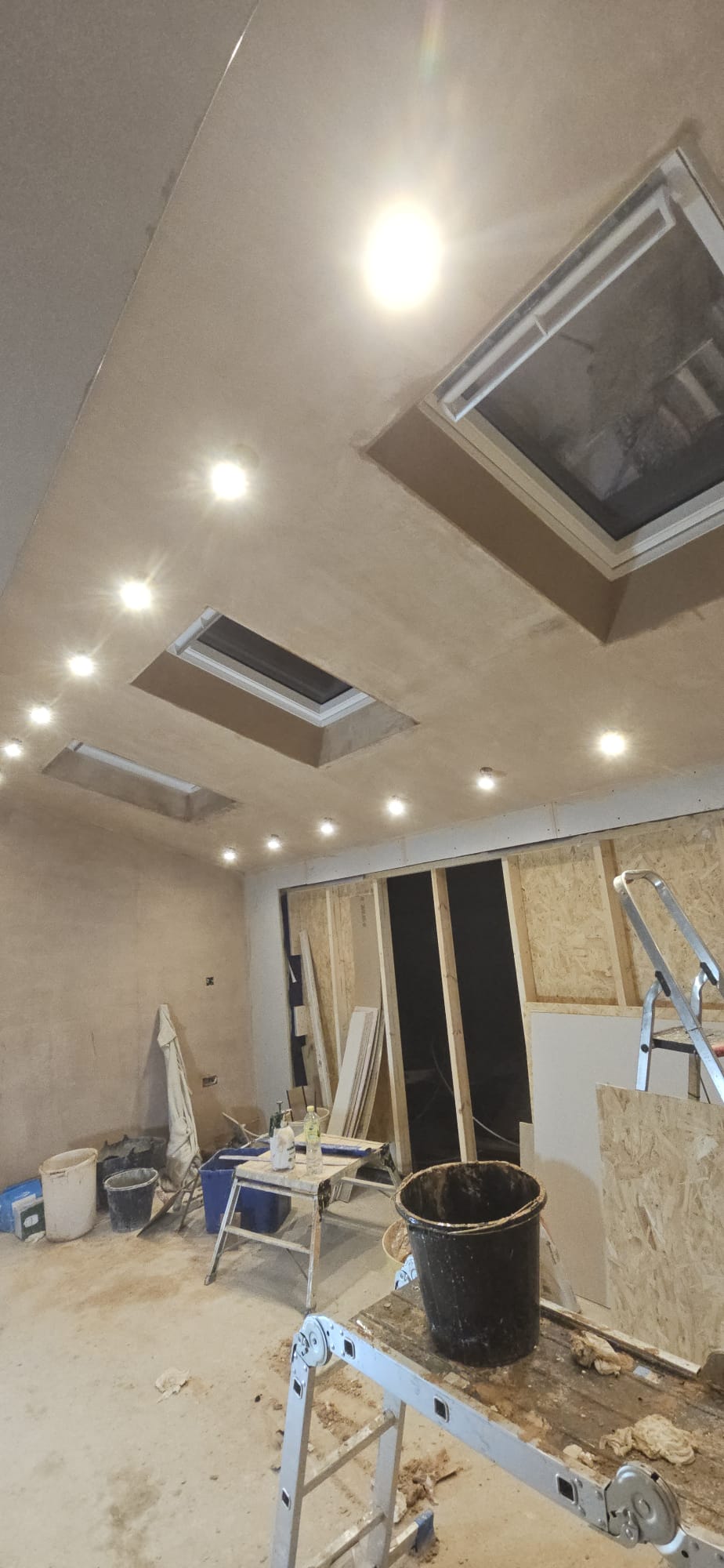Newly plastered sloping ceiling with recessed downlights switched on and three roof windows in an extension under construction